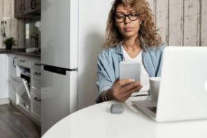 Woman checking her phone seated at table in modern white kitchen working in front of laptop beside fridge and close to wall