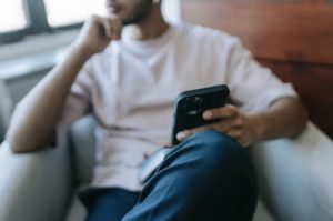 Blurry view of man reclined in chair near window in the background, holding a mobile propped on knee in the foreground