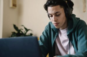 Close up view of casually dressed young man wearing headphones while working on laptop at home