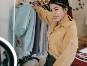 Young woman selecting a robin blue sweater from clothing hanger while engaging with camera in front of ring light