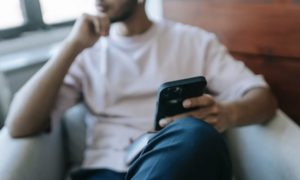 Blurry view of man reclined in chair near window in the background, holding a mobile propped on knee in the foreground