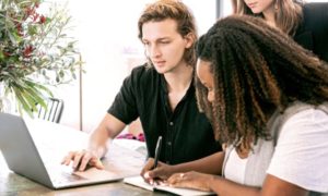 Group of three working from same laptop on table in communal workspace Man clicking through laptop, and woman writing notes