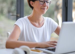 Happy-looking woman in white t-shirt working on laptop in front of window facing greenery outside