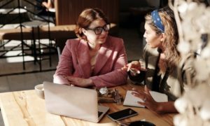 View of two women chatting and working with laptop and taking notes, seated at corner of table in workspace with afternoon light shining on them