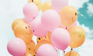 Pink and orange balloons with happy and sad faces floating against a blue and cloudy sky