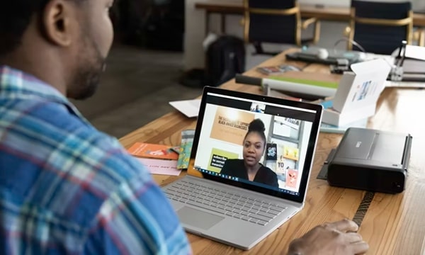 Over the shoulder view of man seated at desk on laptop, chatting with a woman on screen, in messy work area