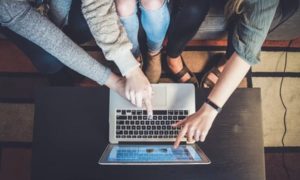 View from ceiling looking down on three women’s arms pointing at open laptop on table