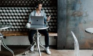 Man working on bench in coffee shop, seated against a geometric backsplash in front of laptop, wearing headphones and checking out smartphone