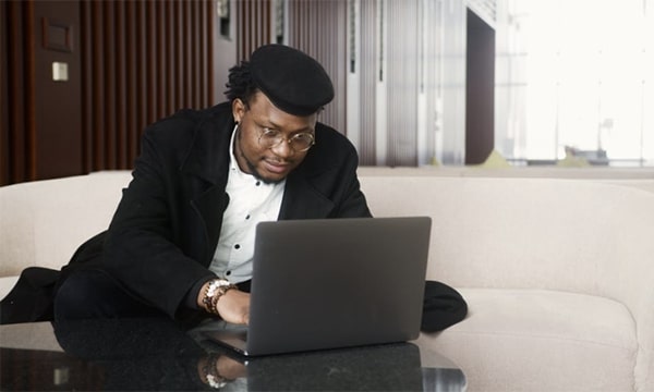 Stylish man wearing a cap, working and seated on a white couch in an open-space, hotel lobby, leaning over and focused on laptop