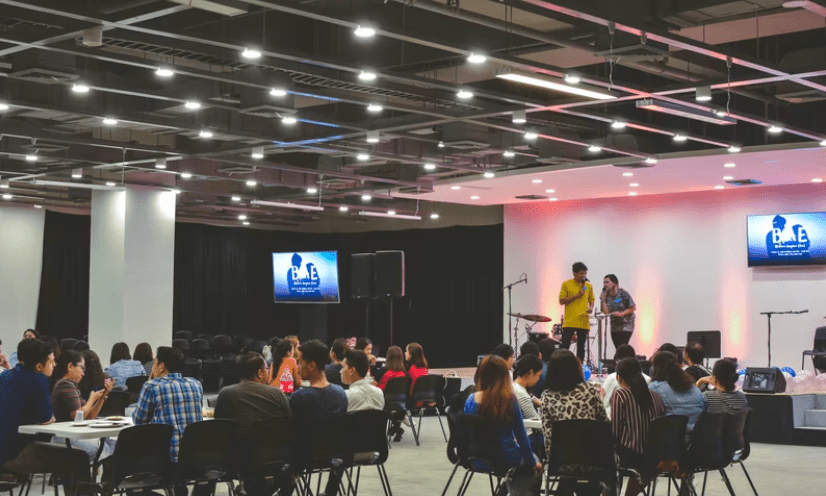 View of hybrid meeting with multiple tables of people, a stage with two hosts, and big screen TVs broadcasting