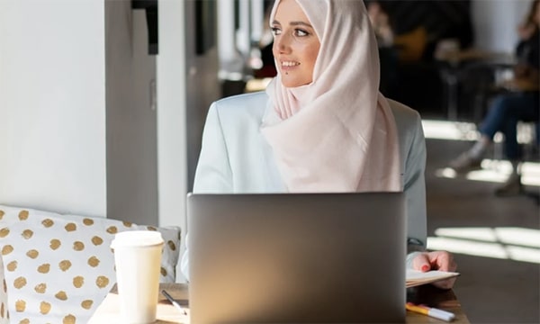 Woman in hijab working on laptop with a take-away coffee, seated in brightly lit coffee shop, looking out the window to the left